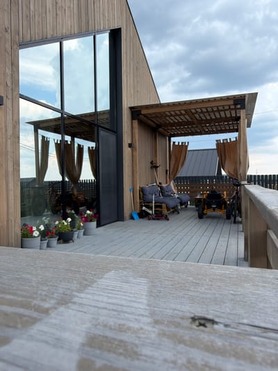 The image shows a wooden deck area of a modern house in the village of Maloye Sedelnikovo, Russia. The house features a large glass window and a wooden exterior. On the deck, there is a covered patio area with two lounge chairs and a small table. Two vehicles, a quad bike and a bicycle, are parked on the deck. Potted flowers are arranged along the edge of the deck. The sky is overcast with clouds. The foreground shows a blurred close-up of a wooden railing.