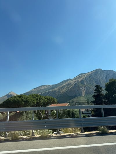 The image shows a view in Cinisi, Italy, looking toward a large mountain under a clear blue sky. The foreground includes a grey metal guardrail and a grey asphalt road with white lines. On the right side of the road is vegetation. Behind the guardrail are trees and houses.
