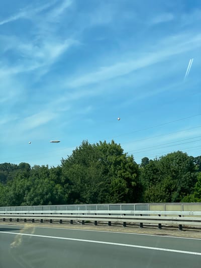 The image depicts a sky with scattered clouds over a wooded area. In the sky, there are several aerial objects. The most prominent is a blimp with the word "GOODYEAR" visible on its side, appearing to be a blimp. To the left of the blimp, there is a spherical object. Further to the left, another elongated, disc-shaped object is visible. In the upper right portion of the image, two parallel contrails from an aircraft are visible against the blue sky. The scene is framed by a highway guardrail and the surface of a road in the foreground. The presence of the Goodyear blimp suggests the location might be near an area where such advertising or transport occurs. Given the detail provided regarding Langenfeld (Rheinland), Germany, and the common presence of blimps for advertising or events, it's plausible this image was taken in the vicinity of Langenfeld, potentially from a highway or elevated vantage point.