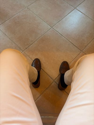 The image depicts a first-person, downward view of a person's lower legs and feet within an interior setting in Cagnes-sur-Mer, France. The individual is wearing light beige trousers, dark blue socks, and brown leather loafer-style shoes. The feet are positioned apart, resting on a floor composed of square, light brown ceramic tiles arranged in a diagonal grid pattern with visible light-colored grout lines. The tiled surface reflects ambient light, indicating an illuminated indoor environment.