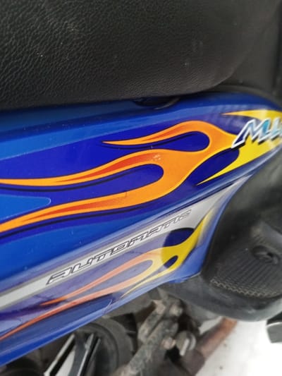 A close-up of a blue motorcycle's side panel shows a vibrant design. Orange flames with black outlines are prominently featured, leading to yellow accents towards the front. "Matic" is printed in a light color along the bottom of the panel, also with flame decorations. The motorcycle seat, covered in black, textured material, is visible in the background. The engine parts and exhaust system can be partially seen below the side panel.