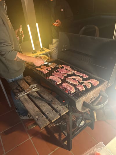 A tilted eye-level shot captures a barbecue scene. A charcoal grill is loaded with grilling meat. A person is using tongs to move the meat. Another person stands nearby. A bottle sits on a table next to them. The grill is placed on a rustic wooden structure. The scene is illuminated by bright lights, highlighting the warmth of the grilling process.