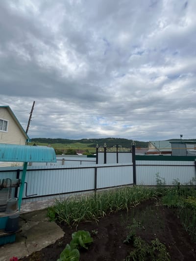 The image depicts a cloudy day in a rural setting, likely in or near Bolshe Kukmor, Russia. The foreground features a garden with rows of green plants, possibly onions or garlic, growing in dark soil. To the left, a portion of a light-colored house with a blue roof is visible, along with a wooden utility pole and overhead wires. A light-colored metal fence runs along the right side of the image, separating the garden from the background.

The background reveals a rolling landscape with green hills and trees. Several buildings, including houses and what appears to be a garage or shed, are scattered throughout the scene. The sky is filled with heavy, gray clouds, suggesting an overcast or approaching rain. The overall impression is one of a quiet, utilitarian rural property on a cloudy day. There is no visible text in the image. The scene is composed of natural elements like clouds, hills, and vegetation, as well as man-made structures like houses, fences, and utility poles. No people or animals are clearly visible.