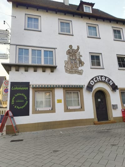 The image shows a white building with multiple windows, located in Neuenstadt am Kocher, Germany. The building has a sign that says "Metzgerei Lohmann" which translates to "Butcher Lohmann" and also "Eigene Schlachtung & Partyservice," indicating that they butcher their own meat and offer party services. Another sign above the entrance arch reads "OCHSEN," which means "Oxen" or "Bullocks." A mural depicting a man holding a goblet and grapes is painted on the building's facade, with the text "Vesper-Stüble" beneath it, likely indicating a place to eat or drink. There are several traffic signs visible on the left side of the image, including a speed limit sign of 20 and a parking prohibition sign. The building appears to be a business with living quarters above. The area in front of the building is paved with cobblestones, and there are some red barriers and a small trash can. The sky is overcast.