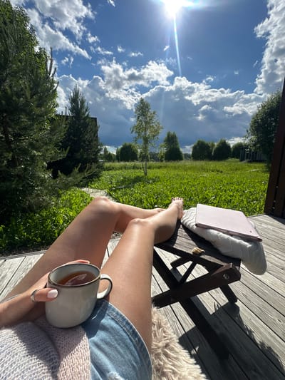 The image depicts a first-person perspective of a person relaxing outdoors on a wooden deck. The person is holding a mug of tea in their left hand and wearing denim shorts and a light-colored knitted cardigan. Their legs are extended, with one foot resting on a folded blanket beside a pink tablet. The deck overlooks a lush green lawn with scattered trees and a few modern houses in the background. The sky is blue with scattered white clouds, and the sun is shining brightly, casting shadows on the deck. The overall scene suggests a peaceful and leisurely moment in a suburban or rural setting. The location is specified as Znamenka, Russia.