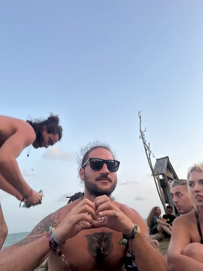 This image depicts an outdoor gathering in Ban Pang Pring, Thailand, under a clear, light-blue sky with faint clouds, suggesting late afternoon or early evening. In the immediate foreground, a shirtless male with dark curly hair and a beard, wearing black sunglasses, is centered. He has multiple festival wristbands on both wrists, a prominent chest tattoo resembling an animal head, a silver chain with a black pendant, and a ring on his right ring finger. His hands are positioned in front of him, forming a heart shape. To his left and slightly behind him, another shirtless male with dark hair tied in a bun is blurred, bent over, and appears to be handling a small object. To the right of the central figure, a partial view of a blonde female wearing a black top and a male with short hair are visible, both looking generally towards the right side of the frame. Further in the mid-ground, several indistinct individuals are seated on the ground. The background features a bare, multi-branched tree-like structure with small lights strung along it, positioned near a weathered, angled wooden structure. A faint horizon line indicating a body of water is visible in the distant background. The overall scene suggests a casual, social event or festival by the water.