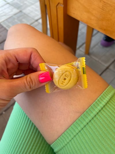 In Plodopitomnik, Russia, a person with bright pink fingernails holds a small, individually wrapped yellow cookie with the letters "H E" embossed on it. The cookie is in clear plastic packaging. The person is seated on a wooden chair, with their legs visible. They are wearing a lime green ribbed top. The background suggests an outdoor setting, possibly a patio or garden, with patterned pavement and wooden furniture.