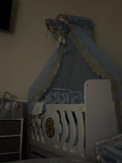 The scene depicts a dimly lit bedroom, specifically a nursery area, located in Preston, United Kingdom. Central to the image is a white wooden baby crib, featuring blue interior padding made of crushed velvet. A light blue sheer canopy, adorned with gold lace trim and a top border of blue fabric roses, hangs over the crib. Small white cloud-like or animal-shaped decorations dangle from the canopy's framework. The crib's visible side panel is white with blue accents and includes a circular, ornate gold medallion. To the left of the crib, a white dresser with three dark grey drawers is visible. On top of the dresser, a grey fabric caddy contains baby care items, including bottles and brushes. A dark, wall-mounted television or screen is partially visible on the upper left wall. No individuals are present, and no actions or interactions are occurring.