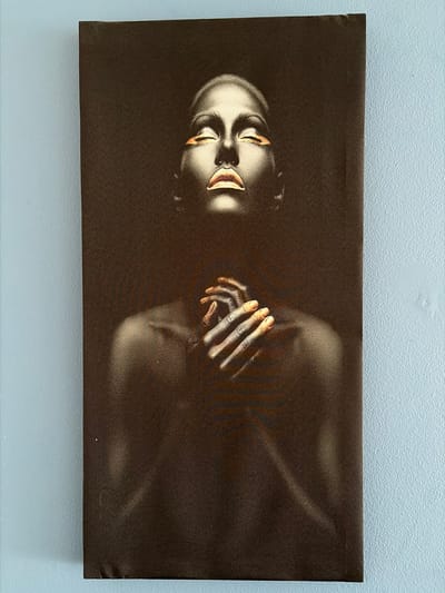 A horizontal rectangular canvas print depicting a person with dark skin, gold accents on their lips and eyelids, and hands clasped near their face, is mounted on a light blue wall. The background of the print is dark brown, fading to black around the edges. The person appears to be floating or lying in a dark space. There is no indication of Lørenskog, Norway.