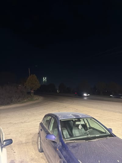 At night in Agigea, Romania, a dark blue hatchback vehicle, possibly an Opel Astra H, is parked on a light-colored, unpaved or dusty surface in the foreground, facing towards the right. The vehicle's hood and roof are visibly coated with a layer of dust or pollen. The interior shows dark-colored seats, a steering wheel, and a hanging air freshener. A portion of a white vehicle is visible parked to the left of the blue car.

In the mid-ground, a paved road, likely a highway, runs diagonally from left to right. Two vehicles are in motion on this road, indicated by blurred headlights on the left and a blurred shape with a red light (possibly taillight) further right, suggesting traffic flow. A smaller road or turn-off curves from the left towards the main road. Sparse vegetation, including a small tree, lines the immediate area and the main road. A utility pole is situated on the left mid-ground.

In the far background, the prominent, illuminated structure of the Agigea Bridge over the Danube-Black Sea Canal is visible, characterized by multiple bright vertical light sources corresponding to its pylons. The sky above is dark. No individuals are visible.