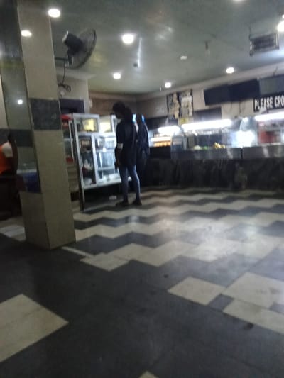 The image shows the interior of a restaurant or cafeteria. The scene is dimly lit, with the only light source being the overhead fluorescent lights. Two people are visible, both wearing dark clothing and standing near a glass display case containing food. The display case is behind a counter with a sign that reads "PLEASE CROSS." The floor is tiled with a checkered pattern of black and white squares. There is a large pillar on the left side of the image. The image was taken in Ovwian, Nigeria, at coordinates 5.503 latitude and 5.799 longitude.
