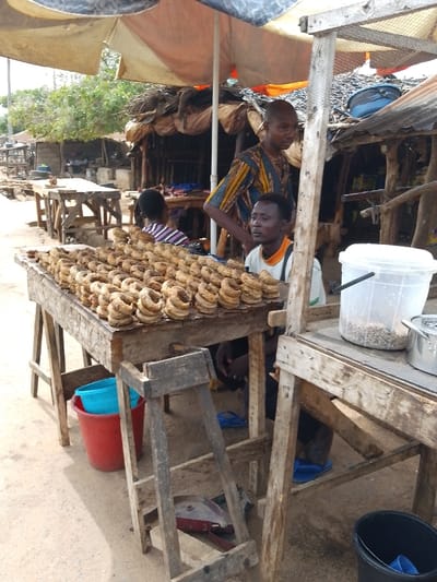 The image depicts an outdoor market stall in Igbo Ijaiye, Nigeria. In the foreground, a rustic wooden table is laden with numerous light-brown, ring-shaped food items, stacked in multiple rows. To the right, a second wooden table holds a clear plastic bucket containing light-colored grains or legumes, and a small metal pot.

Three individuals are present. A bald adult male, wearing a patterned blue, yellow, and brown tunic, stands behind the main display table, facing slightly left. Below and to his right, a second adult male with short dark hair is seated or crouching, wearing a light-colored t-shirt with orange trim, looking to the right. A third individual, seen from behind, wearing a striped shirt, is partially visible in the middle ground, further back between the two men.

The scene is shaded by an overhead, light-colored umbrella canopy. The ground consists of compacted dirt. In the background, several rudimentary market structures constructed from wooden poles and corrugated metal sheets are visible, with foliage appearing beyond them. Various objects are on the ground, including a red bucket containing a light blue basin, a dark blue bucket, and several pairs of blue flip-flops. A small red and black object lies beneath the main display table.