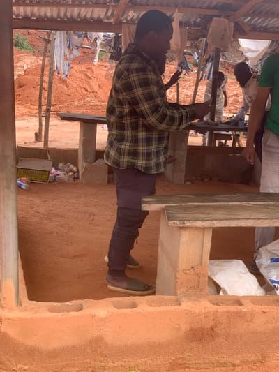 The image depicts an outdoor scene under a rustic, open-air shelter with a corrugated metal roof. The ground is covered in reddish-brown earth. In the foreground, a man wearing a plaid shirt and dark pants is standing, holding a small object in his hands and appearing to interact with it. To his left, a partially opened cardboard box labeled "Black Wood Filters" is visible, along with several empty plastic water bottles.

In the background, the environment suggests a rural or undeveloped area, with piles of red earth, possibly from construction or mining. Several other individuals are present in the background, partially obscured by the shelter's structure and other people. One person is wearing a white shirt and dark pants, another is wearing a striped garment, and another person is wearing a green shirt. The scene appears to be a market or a gathering point. The overall impression is one of an informal, everyday setting in a somewhat dusty environment. The presence of the "Black Wood Filters" box and plastic bottles suggests commercial activity or waste accumulation.