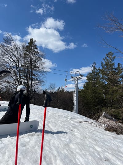 A partially snow-covered ski slope in Bogoslov, Bulgaria, is depicted under a blue sky with scattered white cumulus clouds. In the foreground, a person clad in black winter outerwear and a white helmet is seated on a white snowboard, positioned with the snowboard base on the snow. Two red ski poles with black grips are planted upright in the snow to the right of the person. A tall, gray metal ski lift pylon, equipped with a ladder on its side, stands prominently in the midground, supporting a cable from which multiple ski chairs are suspended; one chair features a red safety component. The background consists of a dense forest with a mix of bare deciduous trees and green coniferous trees, extending across the hillside. The ground cover is primarily snow, with some patches of exposed dirt and brush visible near the edge of the tree line.