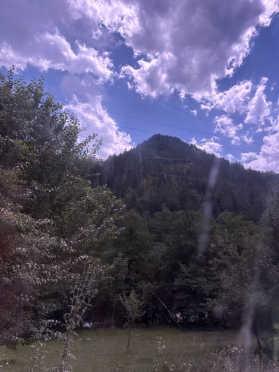 The image shows a mountainous landscape with a dense forest covering the slopes. The sky is blue with scattered white clouds. There are power lines running horizontally across the frame, intersecting with the trees and the mountain. In the foreground, there is a grassy area with some scattered vegetation. The overall impression is of a natural, undeveloped area. The image does not provide any specific visual cues to suggest it is located in Aziatskii, Russia.