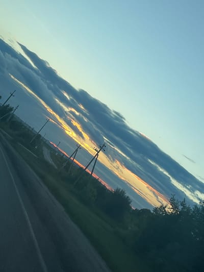 A diagonal view of a rural road in Kivsertkasy, Russia, at sunset. The sky is a gradient of light blue with long, dark clouds tinged with orange and yellow from the setting sun. Power lines with utility poles are visible along the side of the road, traversing the landscape. The road itself is paved and appears to be empty. Green vegetation, including trees and grass, lines the sides of the road, with the foliage casting shadows in the fading light.