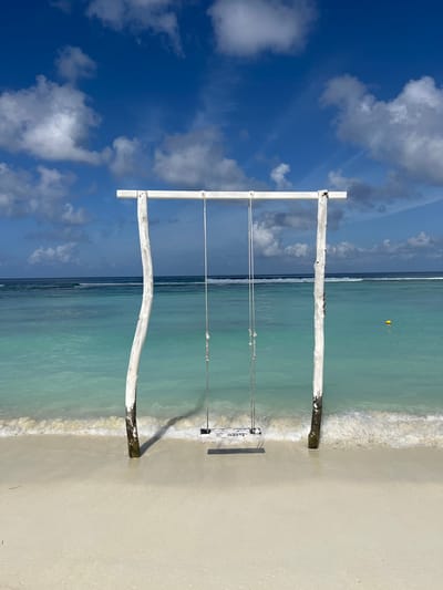 A wooden swing set with a white horizontal bar and two white, gnarled posts is positioned on a sandy beach. The swing seat is made of weathered white wood. The turquoise ocean stretches out behind the swing set, with gentle waves lapping at the shore. In the distance, the horizon is visible, where the ocean meets a bright blue sky filled with white, fluffy clouds. A yellow buoy floats on the water to the right of the swing set. The scene appears to be in the city of Malé, Maldives.