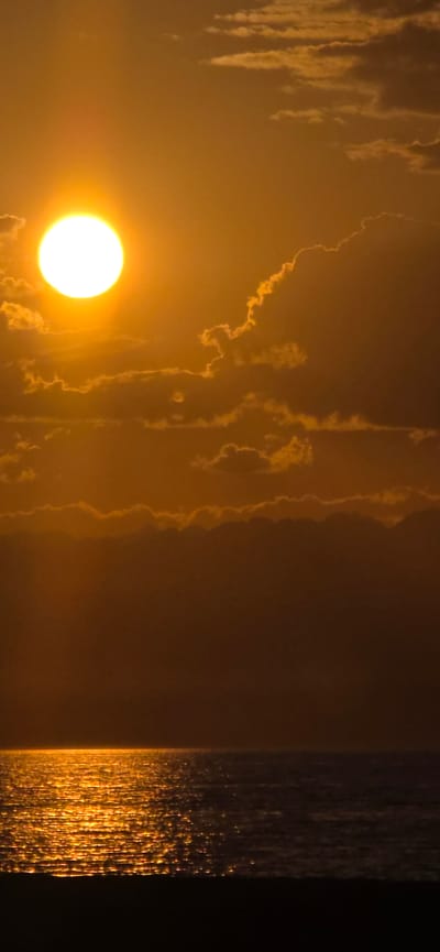 This is a vertical, full-shot photograph of a sunset over the sea in Boz Beshik, Kyrgyzstan. The sun, a bright white orb, is positioned in the upper left quadrant, casting a warm golden glow across the sky and the rippling water. Wispy clouds, illuminated by the setting sun, dot the sky. The lower portion of the image features a dark silhouette of land, likely a beach or coastline, bordering the shimmering sea.