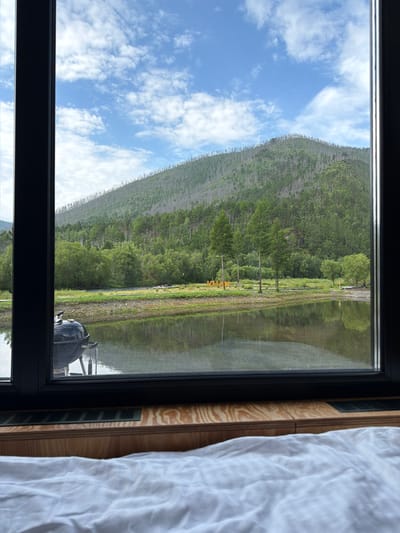 A wide-angle view from indoors looks out through a large window to a serene natural landscape in Bol'shoe Goloustnoe, Russia. In the foreground, a white, crumpled bedsheet is visible on a wooden surface, suggesting an interior space. The window frames a tranquil scene featuring a still body of water that mirrors the surrounding environment.
Dominating the middle ground is a lush green embankment along the water's edge, teeming with various trees and vegetation. Beyond this, a majestic, treed mountain rises, its upper slopes appearing sparse with a noticeable presence of dead trees. The sky above is a vibrant blue, dotted with scattered white clouds.
Visible in the middle ground, on the embankment, are some yellow structures, possibly a sign or art installation, and a black barbecue grill is positioned to the left, near the water. The overall atmosphere is peaceful and picturesque, showcasing the natural beauty of the region.