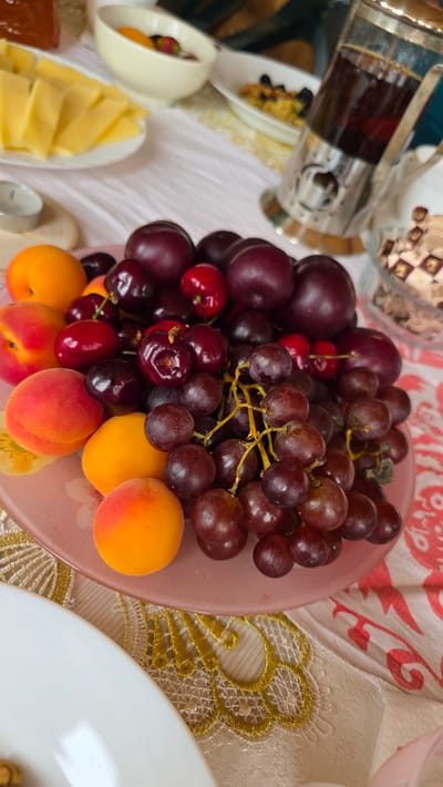 A close-up, eye-level shot captures a platter of fresh fruit on a table covered with a pink and white patterned tablecloth. The platter, made of pink glass, is filled with a vibrant assortment of red grapes, dark red cherries, and orange apricots. The fruits are arranged in a natural, appealing way, suggesting a leisurely meal or snack. In the background, slightly out of focus, are other elements of a meal setting. To the left, a white plate holds sliced yellow cheese. Further back, a white bowl contains a mix of fruits, and a clear glass French press with dark liquid stands to the right. The scene is brightly lit, possibly by natural light, highlighting the textures and colors of the fruits. The overall impression is one of abundance and a healthy, fresh offering. The exact location in the city of Купоярово, Russia, cannot be determined from the visual information provided.