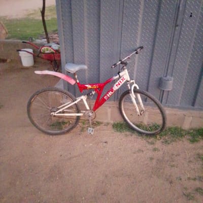 A red and white Phoenix brand bicycle with suspension is parked on a dirt and gravel surface in front of a corrugated metal wall. The bicycle has a light-colored seat, white handlebars, and white front suspension forks. The frame is predominantly red with "PHOENIX" written in white text along the lower tube and "SPORT" written on a smaller tube. The rear tire is on the left side of the image and the front tire on the right. A red wheelbarrow with a white bucket and green objects is visible in the background to the left. The environment appears to be an outdoor area, possibly a yard or an open space in Wuro Allahamdu, Nigeria, with some sparse green vegetation visible.