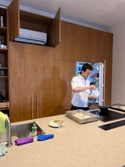 The image depicts a modern kitchen interior located in Agios Ioannis Diakoftis, Greece. The dominant feature is a wall entirely clad in light-brown wood veneer cabinetry, extending from floor to ceiling. An integrated Hitachi air conditioning unit is visible within an open upper cabinet on the left side of this wall, with two cabinet doors above it also open upwards. Below the air conditioner and extending to the right are numerous closed cabinet doors. On the right side of the main wall, an integrated refrigerator is open, revealing various items including canned beverages and bottled water.
A single male individual, a young adult with dark hair, is positioned in front of the open refrigerator. He wears a white, textured short-sleeved collared shirt and dark trousers. His attention is directed downwards as he holds a white rectangular container in his left hand and appears to be pouring or transferring contents from a smaller, white disposable container held in his right hand.
In the foreground, a light-colored, speckled countertop spans the width of the frame. On the left, a stainless steel sink features a chrome faucet, a green pump-bottle soap dispenser, a red cleaning scrubber, and three microfiber cloths in purple, blue, and light green. To the right of the sink, a small white plate holds two halves of a passion fruit, partially covered with clear plastic wrap. Further right, a black induction cooktop with four burners is visible. A small black saucepan rests on one of the cooktop burners. Adjacent to the cooktop, a large, rectangular silver-colored baking pan is placed on the countertop.