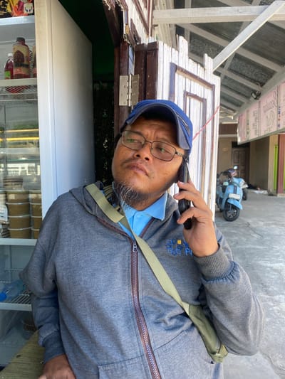 A medium shot captures a man with glasses and a blue cap holding a black smartphone to his right ear. He is wearing a grey zip-up hoodie over a blue polo shirt and a khaki-colored crossbody bag. The man is standing in front of an open doorway with a white wooden doorframe and a weathered, possibly white, paneled wall. Behind him, a glass-fronted refrigerator with various products is visible on the left, and a glimpse of outdoor space with a light blue scooter and what appears to be a person lying on the ground in the background is seen through the doorway on the right. The environment suggests a commercial or street-side setting, possibly in Sungai Paring, Indonesia, with the corrugated metal roof and wooden structures indicating a casual, possibly tropical, location. The lighting suggests it is daytime.
