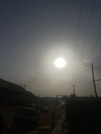 The image captures a street view in Unguwar Fatika, Nigeria, under bright sunlight. Power lines traverse the sky, connected to utility poles. Buildings line the street, with parked vehicles visible. A person is walking along the sidewalk.