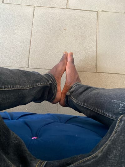 The image, captured in Igbesa, Nigeria, presents a close-up, first-person view of an individual's lower legs and bare feet while seated. The subject is wearing dark, faded denim jeans. Both bare feet are positioned together on a light grey tiled floor, with the soles pressed against each other and the heels slightly raised, forming a distinct upward-pointing V-shape. The tiled floor features rectangular tiles with visible grout lines and minor surface marks. Directly beneath the denim-clad legs, a portion of a blue, upholstered surface, possibly a tufted cushion or seat, is visible. A small pink object is discernible within one of the tufted depressions of the blue upholstery.