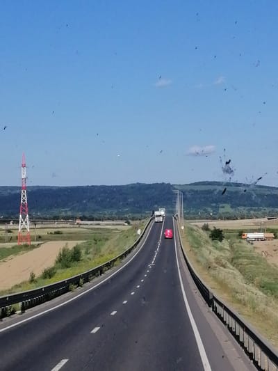 The image depicts a highway in a rural area. A bright red and white tower, possibly for electrical transmission, stands to the left of the road. A red van and a white truck are visible on the highway, traveling in the same direction. The road is bordered by grassy slopes and fields. In the background, rolling hills covered in greenery recede into the distance under a clear blue sky. The scene appears to be a bright, sunny day. There are no obvious indications of the city of Nicolae Bălcescu, Romania, within the image.