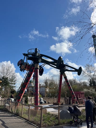 The image displays an outdoor scene within an amusement park in De Panne, Belgium, under a clear blue sky with scattered white cumulus clouds. The central subject is a large mechanical amusement ride, likely an inverted pendulum or giant swing attraction. This ride features a robust, dark-colored metal superstructure from which a long arm extends, currently positioned with passenger gondolas on the left side. The primary support columns of the ride are adorned with a distinctive red and dark green/black diamond or zigzag pattern. Multiple passengers are visible within the dark red and brown seats of the gondolas on the left side of the arm. Ladders are affixed to the ride's support columns.
In the background, themed amusement park architecture, characterized by white buildings with green accents, domes, and spires, is visible. Trees with bare or minimal foliage are present across the mid-ground and background, particularly along the right edge. The foreground features a paved pathway on the left, separated from a landscaped area by a low barrier constructed from wooden posts and rope netting. This landscaped area contains artificial rocks and sparse ground cover.
On the paved pathway in the lower right, an adult figure, seen from the back in a dark jacket and pants, stands next to a dark-colored, four-wheeled baby stroller. A second individual is partially visible behind them to the right. Additional park visitors are observable in the mid-ground behind the barrier, near the base of the ride, either standing or moving.