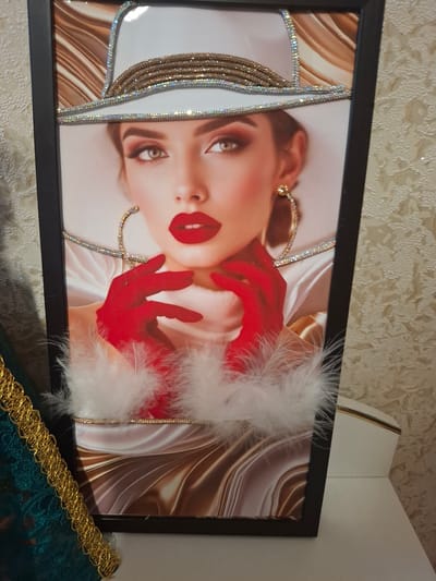This is a framed artwork featuring a close-up of a woman's face. The woman has striking makeup, with bold red lips and dramatic eye makeup. She is wearing a white hat adorned with rhinestones and gold trim. Her hands are also covered in red gloves, and she is surrounded by white feathers. The background of the artwork is abstract, with swirling patterns of brown and gold. The artwork is displayed in a dark frame and is positioned against a textured, light-colored wall. A portion of a decorative gold trim is visible in the foreground. The overall impression is one of glamour and artistry. The specific location of Jitiqara, Kazakhstan, is noted, but there are no visual cues within the image to confirm or suggest this location. No text is visible in the image.