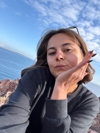 A close-up, selfie-style photograph captures a young adult female outdoors in Théoule-sur-Mer, France. The subject, with medium-length brown hair, dark eyes, and a fair complexion, is wearing a black long-sleeved crew-neck top. Sunglasses are positioned on top of her head. Her left hand, adorned with dark nail polish on elongated fingernails and a small ring on the ring finger, rests against her chin and jawline. Her lips are subtly pursed, and she gazes directly into the camera. Bright sunlight illuminates her face from the right. The background features a clear blue sky with scattered white cumulus clouds, above an expansive view of the deep blue Mediterranean Sea. Reddish-brown rocky terrain, characteristic of the Esterel Massif, is visible in the lower left foreground. A yellow buoy is discernible in the distant water, and a hazy coastline stretches along the far horizon.