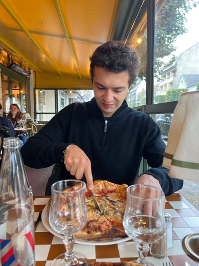 A young adult male is seated at a checkered table on a restaurant terrace in Deauville, France, actively cutting a pizza. He wears a black quarter-zip long-sleeve top and uses a knife to portion a pizza featuring red sauce, cheese, and dark toppings on a white plate. Two empty stemmed glasses, a clear glass bottle with a red, white, and blue label, and a salt shaker are on the table. In the background, another female diner is visible at a separate table. The terrace is covered by a yellow awning with a black radiant heater mounted beneath it. Beyond the glass partitions of the establishment, trees and residential or commercial buildings are discernible under an overcast sky. A beige lampshade with green stripes is partially visible on the right side of the frame.