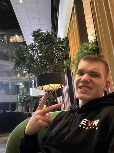 A person wearing a black hoodie with "EVN SPORTS" printed on it is making a peace sign. They are sitting indoors with plants and a circular lamp in the background. Outside the window are several floors. The image is rotated counter-clockwise approximately 90 degrees. The person is smiling and looking at the camera.