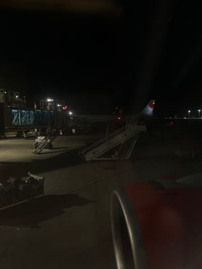 An airplane sits on a dark tarmac at night, connected to a passenger boarding bridge. A portable stairway is positioned near the plane's tail. The tail features a red and white design. The area is illuminated by lights from the boarding bridge and nearby ground equipment. The aircraft is viewed from a high angle, possibly from inside another airplane. An engine cowling and a metallic ring are visible in the foreground.