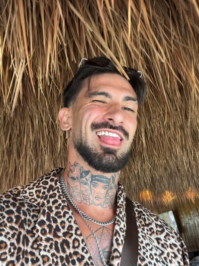 A smiling man with a beard and tattoos on his neck and chest is lying on a bed of dried grass. He is wearing a leopard print shirt and a silver chain necklace. Sunglasses are propped on his head. The scene is at an undetermined location in Ban Khlong Khian บ้านคลองเคียน, Thailand.