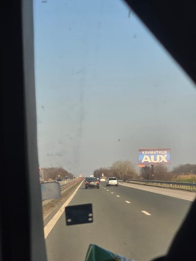 The image presents a daytime view from inside a vehicle, looking forward through a smudged windshield onto a multi-lane highway. Dark framing from the vehicle's interior partially obstructs the left, top-right, and bottom-right sections of the frame. The sky is clear and blue.
The highway is asphalt, featuring at least two lanes of travel in the depicted direction, separated by dashed white lines. A solid white line borders the right shoulder. A central median barrier, composed of a metal guardrail atop a raised strip, separates the opposing lanes of traffic.
Several vehicles are visible ahead on the highway. A dark-colored sedan is in the left lane, followed by a white car in the right lane further ahead. Other vehicles are present in the distance.
On the right side of the highway, beyond a metal guardrail, a large billboard is erected on two poles. The billboard displays text in Bulgarian: "КЛИМАТИЦИ" (Air Conditioners) in white on a blue background, with "AUX" in large white letters on a red-to-blue gradient below it. The surrounding environment features bare deciduous trees and shrubs, indicative of late winter or early spring, extending into a field or open area. Similar bare trees are visible beyond the median on the left.
Within the camera vehicle's interior, a small, black, rectangular object with four circular perforations (two top, two bottom) is visible on the windshield, possibly an attachment or sensor. A green object, possibly part of packaging, is partially visible in the bottom-right corner.
The scene is located on a highway in or around Trilistnik, Plovdiv Province, Bulgaria.