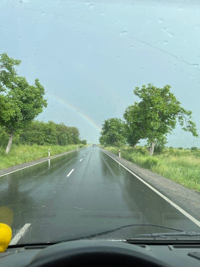The image shows a view from inside a vehicle, looking out onto a wet asphalt road stretching into the distance. The perspective is slightly angled. There are white lane markings on the road. Trees line both sides of the road. A rainbow is visible in the sky. The image was taken while driving through the city of Pevtsite, Bulgaria.