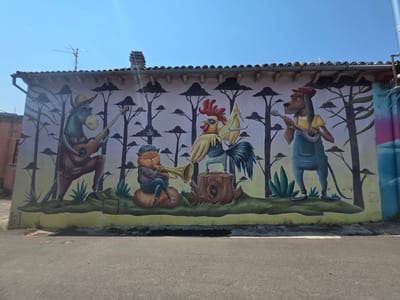 This is a wide-angle photograph of a mural on the exterior wall of a building, likely in Viterbo, Italy, given the context. The mural depicts a band of anthropomorphic animals playing instruments against a stylized backdrop of trees and a clear blue sky.
From left to right, the mural features:
* A donkey wearing a hat and playing a guitar.
* A cat-like creature in a hat and vest, sitting on a rounded surface, playing a trumpet.
* A rooster wearing sunglasses, standing on a tree stump, holding a triangle and beater.
* A dog wearing overalls and a hat, playing a ukulele-like instrument.
The backdrop consists of a pale pinkish-purple wall with silhouetted trees with dark, rounded tops. A yellow and green band runs along the bottom of the mural, representing the ground and foliage. The building has a tiled roof with visible eaves and a chimney. Antennas are present on the roof. The photograph is taken from a street-level perspective, showing the asphalt road in the foreground. There are some graffiti tags on the far right side of the mural. The lighting suggests it is daytime, with bright sunlight casting some shadows.
