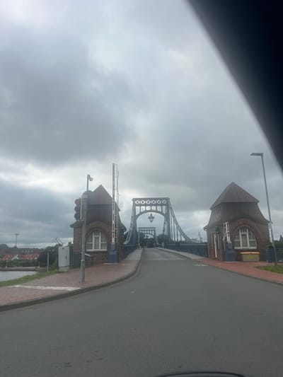 This image shows a view from a vehicle looking out at a bridge and its surroundings on a cloudy day. The primary subjects are the distinctive brick structures of what appear to be bridge toll booths or control houses, a large suspension bridge in the background, and a paved road in the foreground. Two brick buildings with arched windows and dark, pointed roofs are prominently featured on the right side of the frame. They have white barriers and traffic signals, indicating a functional crossing.
The bridge itself is a significant structure with intricate metal latticework and suspension cables, suggesting it is an older, perhaps historical, bridge. The sky is overcast with dense grey clouds, indicating a dull or potentially rainy day. The road curves away from the viewer, with a brick-patterned sidewalk beside it. In the far background, more buildings and what looks like a construction crane can be seen, hinting at an urban or industrial setting near a body of water.
There is some yellow graffiti on one of the buildings, but it is difficult to read clearly. The overall mood of the image is somber due to the weather, but the architectural elements of the bridge and buildings suggest a place of historical or engineering significance. The scene captures a moment of transit, possibly as the vehicle is approaching or leaving the bridge. The location context of Wilhelmshaven, Germany, aligns with the visual cues of a port city with significant infrastructure.