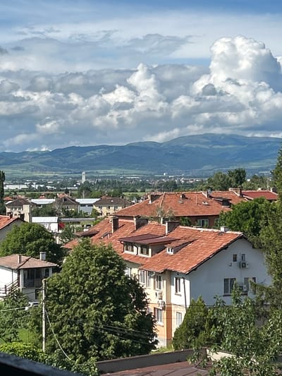 This panoramic image presents a wide-angle view from an elevated perspective, overlooking the town of Gara Elin Pelin, Bulgaria, nestled within a valley and framed by distant mountains. The scene is captured on a bright, partially cloudy day.
In the foreground, the view is dominated by the reddish-brown tiled roofs of numerous residential buildings, interspersed with the lush green canopies of mature trees. Many buildings have light-colored facades, and several air conditioning units are visible on their exterior walls. Utility poles and overhead wires crisscross some sections.
The mid-ground reveals the broader expanse of the valley, with more scattered buildings, cultivated fields, and possibly roads extending into the distance. A notable detail in the mid-ground is a slender, white tower-like structure rising above the other buildings.
Stretching across the background is a long, undulating range of mountains, appearing soft blue and green under the daylight, indicating their distance. The sky above is a vibrant blue, filled with dramatic, voluminous cumulus clouds that are brightly lit by the sun, creating a dynamic and picturesque overhead canopy.
There are no visible people or specific activities taking place, rendering the scene calm and static. No legible text is discernible within the image. The image captures a serene blend of urban settlement and natural landscape under a clear sky.