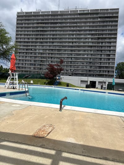 The image depicts a swimming pool complex in Philadelphia, United States, adjacent to a tall, gray, multi-story residential building. The pool is filled with blue water and has a white border and blue tiles. A metal pipe runs along the edge of the pool. A lifeguard chair with an orange umbrella is positioned at one end. The building has many windows and appears to be an apartment building. The sky is cloudy.