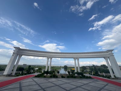 The image captures a wide-angle view of an entranceway featuring a grand, ornate arch supported by Corinthian columns. A vibrant red carpet is laid out on the patterned stone ground, leading towards the arch. Beyond the arch, a fountain sprays water in the center of a landscaped area with manicured bushes and trees. The background reveals a rural landscape with rolling hills and mountains under a bright blue sky dotted with white clouds. The location is identified as the city of Mkhchyan, Armenia. There are no people visible in the image.