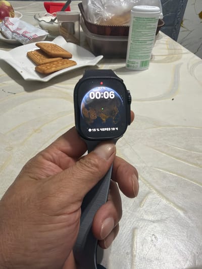 A close-up shot shows a person's hand holding a smartwatch. The smartwatch displays the time "00:06" and "87%". A small green dot is visible on the screen, and text in Russian reads "15% ЧЕРЕЗ 19 Ч". The background is a table with a patterned surface and some objects, including a white plate with three rectangular cookies, a plastic bottle with green text, and a red object that could be a piece of fruit or a toy. The overall setting appears to be indoors, likely a home environment in Aleksandrovka, Kyrgyzstan. The lighting suggests it could be nighttime or a dimly lit room, given the time displayed on the watch. No specific emotions are conveyed by the image.