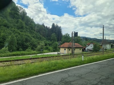 The image shows a rural landscape with a railway track running horizontally across the foreground. On the left side of the track, a steep, tree-covered hill rises. To the right of the track, there is a small yellow building with a red-tiled roof, and further to the right, two houses with tiled roofs are visible. Beyond the houses, a line of trees forms the horizon. The sky is partly cloudy with patches of blue. A signpost reading "347 0" is visible near the houses. The scene appears to be in Agsdorf, Austria, given the context provided. There are no people or animals visible in the image. The overall impression is one of a quiet, natural setting with residential elements adjacent to the railway.