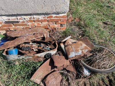 The image shows a patch of overgrown grass in what may be Поедуги, Russia, near a partially collapsed brick wall and concrete base. The scene is littered with rusted metal debris, including what appears to be parts of a bicycle wheel, a metal bucket, and fragmented metal sheets and containers. Electrical wires and components are also visible amidst the debris. The overall impression is one of neglect and decay.