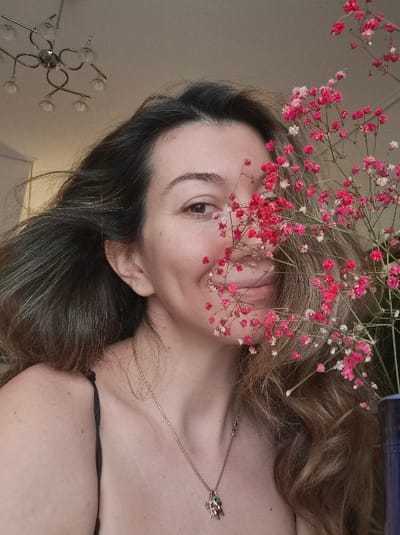The image displays a close-up portrait of a female subject indoors. The subject, with dark brown hair featuring lighter highlights, is positioned in the center-left of the frame, facing slightly right and looking directly at the viewer with a faint smile. Her right eye is fully visible, while the left side of her face, including her left eye, cheek, and part of her mouth, is partially obscured by a dense cluster of small, delicate pink and white flowers, consistent with baby's breath. Her hair is voluminous and falls over her shoulders. She is wearing a dark-colored spaghetti strap top. A thin, gold-colored chain necklace with a small, intricate pendant (possibly an animal figure with green and red accents) rests on her upper chest. In the upper-left background, a silver-colored chandelier with six curved arms, each supporting a glass shade, is suspended from a light-colored ceiling. A segment of a blue object, possibly a vase, is visible on the far right edge of the frame. The exact location within the city of Municipal Unit of Corinth, Greece, cannot be determined from the visual evidence provided in this image.