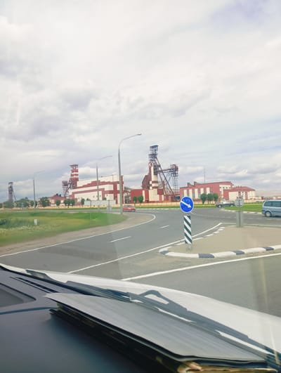 The image depicts a view from inside a vehicle, looking out at a large industrial complex under a cloudy sky. The complex features several tall, red and white structures, likely mine shafts or processing facilities, with distinctive lattice towers. In the foreground, a road curves to the right, with a traffic sign indicating a mandatory direction to the left. A black and white striped pole supports the blue circular sign with a white arrow.
The scene appears to be an operational industrial area, possibly a mining site given the prominent structures. The road is asphalt with white lane markings, and a patch of green grass and some trees are visible on the side. Several vehicles are present, including a red car on the road, a dark-colored SUV, and a light blue van further in the distance. The presence of the vehicles suggests ongoing activity at the location.
The weather appears overcast, with a bright, diffused light that doesn't cast harsh shadows, suggesting it might be daytime, but not necessarily midday. The overall mood is neutral, focused on the industrial landscape and the immediate surroundings of the road. There is no visible text in the image, apart from the symbols on the traffic sign. The location context provided is Краснадворскі сельскі Савет, Belarus, which helps identify the setting as an industrial area within this region.