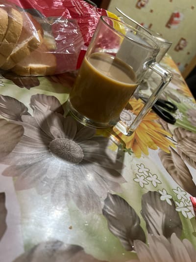 This is a close-up, high-angle shot of a table in Shyshkove, Ukraine. The table is covered with a floral-patterned tablecloth. On the table are several slices of bread in a plastic bag, a glass mug filled with coffee or tea, and a pair of scissors. The lighting is dim, creating shadows and highlights. The overall impression is one of a casual, indoor setting.